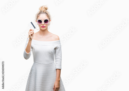 Young beautiful blonde woman holding credit card over isolated background with a confident expression on smart face thinking serious