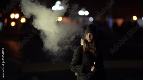 A girl is smoking vape outside in the evening. Nice colorful bokeh in front of the evening city. photo