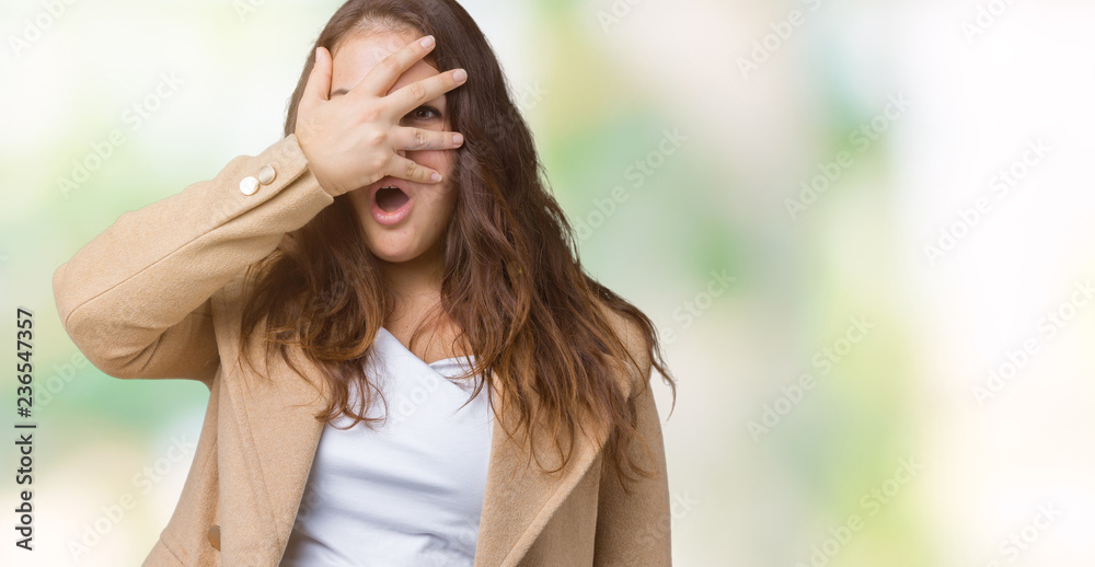 Beautiful plus size young woman wearing winter coat over isolated background peeking in shock covering face and eyes with hand, looking through fingers with embarrassed expression.