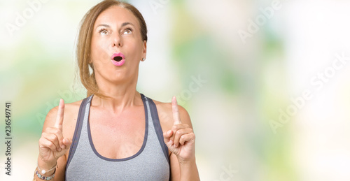 Beautiful middle age woman wearing sport clothes over isolated background amazed and surprised looking up and pointing with fingers and raised arms.