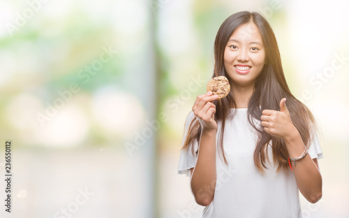 Wallpaper Mural Young asian woman eating chocolate chip cookie over isolated background happy with big smile doing ok sign, thumb up with fingers, excellent sign Torontodigital.ca