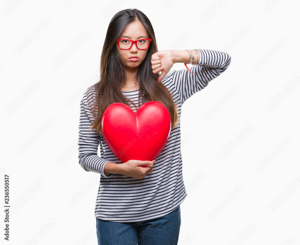 Young asian woman in love holding read heart over isolated background with angry face, negative sign showing dislike with thumbs down, rejection concept Young asian woman in love holding read heart over isolated background with angry face, negative sign showing dislike with thumbs down, rejection concept
