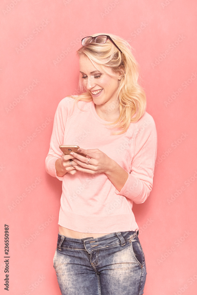 one young woman smiling, looking to camera, while texting or using her smartphone, 20-29 years old, long blond hair. Shot in studio on pink background. one young woman smiling, looking to camera, while texting or using her smartphone, 20-29 years old, long blond hair. Shot in studio on pink background.