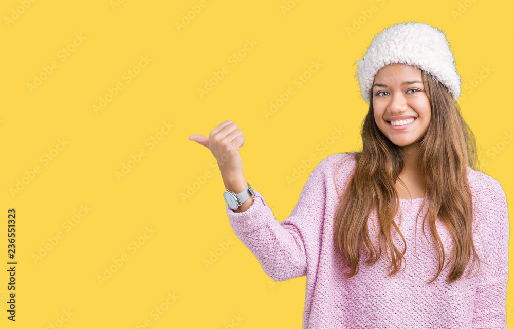 Young beautiful brunette woman wearing sweater and winter hat over isolated background smiling with happy face looking and pointing to the side with thumb up. Young beautiful brunette woman wearing sweater and winter hat over isolated background smiling with happy face looking and pointing to the side with thumb up.