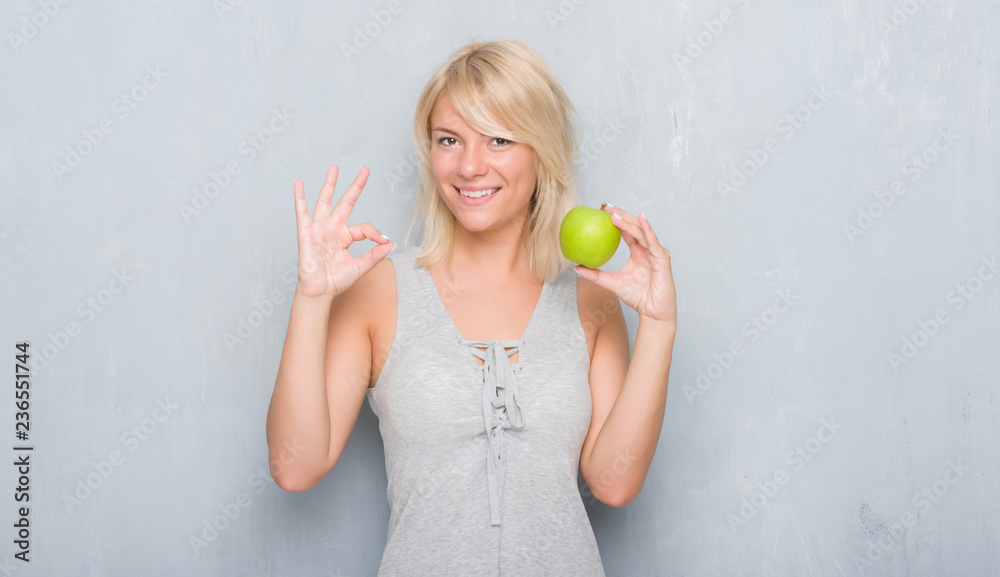 Adult caucasian woman over grunge grey wall eating green apple doing ok sign with fingers, excellent symbol Adult caucasian woman over grunge grey wall eating green apple doing ok sign with fingers, excellent symbol