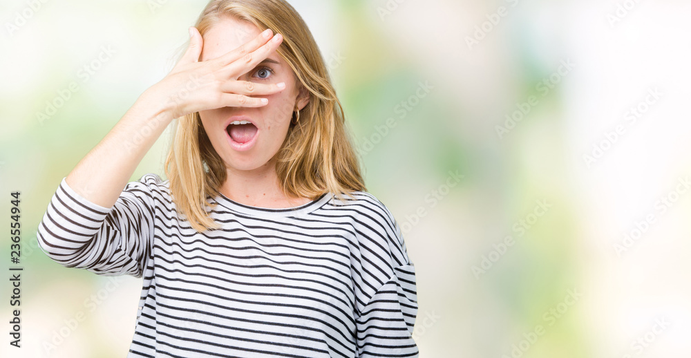 Beautiful young woman wearing stripes sweater over isolated background peeking in shock covering face and eyes with hand, looking through fingers with embarrassed expression.