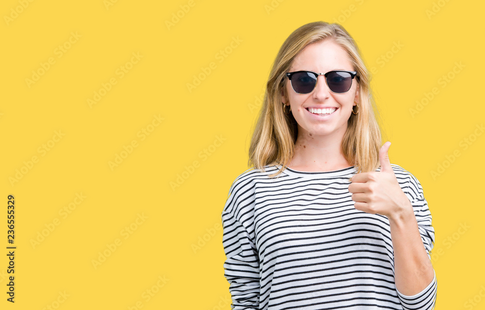 Fototapeta premium Beautiful young woman wearing sunglasses over isolated background doing happy thumbs up gesture with hand. Approving expression looking at the camera with showing success.