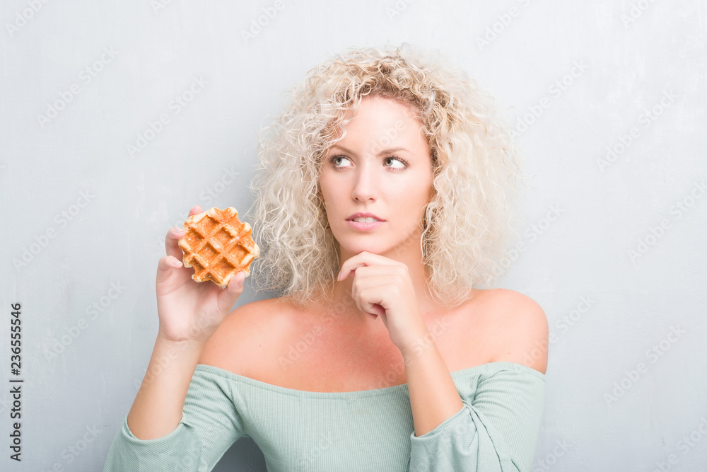 Young blonde woman over grunge grey background eating belgian waffle serious face thinking about question, very confused idea Young blonde woman over grunge grey background eating belgian waffle serious face thinking about question, very confused idea