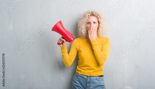 Young blonde woman over grunge grey background holding megaphone cover mouth with hand shocked with shame for mistake, expression of fear, scared in silence, secret concept
