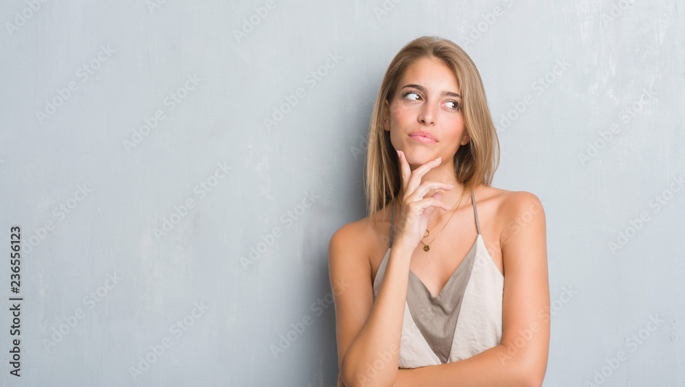 Beautiful young woman standing over grunge grey wall serious face thinking about question, very confused idea Beautiful young woman standing over grunge grey wall serious face thinking about question, very confused idea