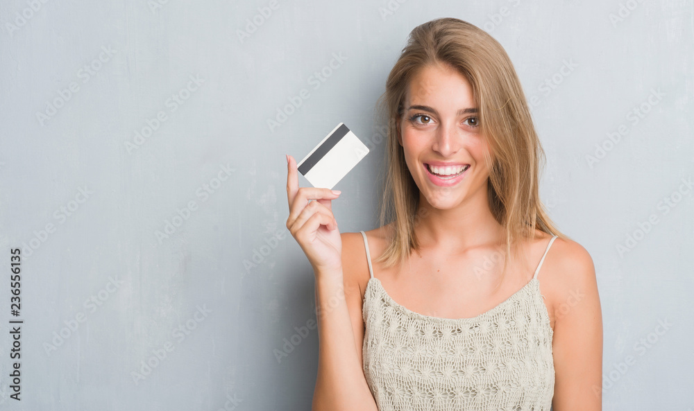 Fototapeta premium Beautiful young woman over grunge grey wall holding credit card with a happy face standing and smiling with a confident smile showing teeth