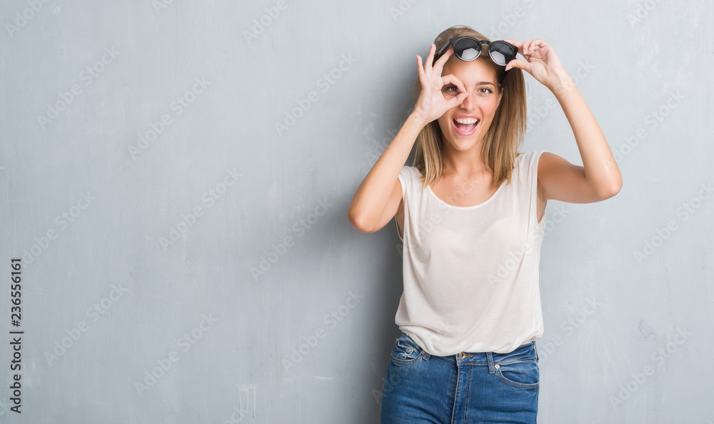 Beautiful young woman standing over grunge grey wall wearing fashion sunglasses with happy face smiling doing ok sign with hand on eye looking through fingers Beautiful young woman standing over grunge grey wall wearing fashion sunglasses with happy face smiling doing ok sign with hand on eye looking through fingers