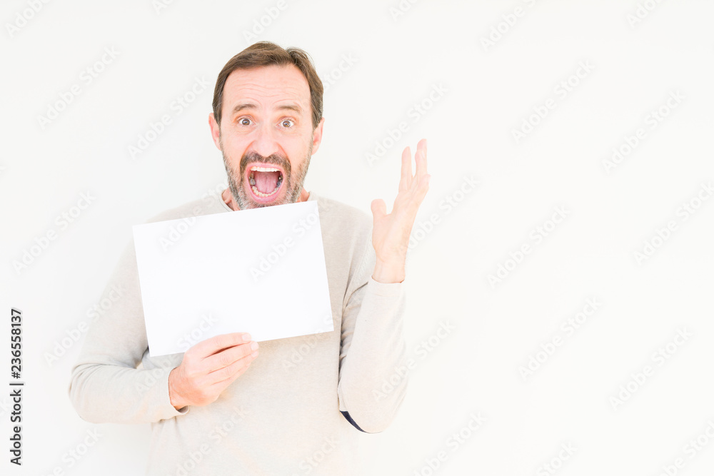 Senior man holding blank paper sheet over isolated background very happy and excited, winner expression celebrating victory screaming with big smile and raised hands Senior man holding blank paper sheet over isolated background very happy and excited, winner expression celebrating victory screaming with big smile and raised hands