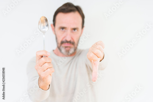 Senior man holding silver spoon over isolated background with angry face, negative sign showing dislike with thumbs down, rejection concept