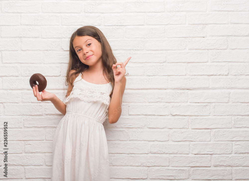 Young hispanic kid over white brick wall eating chocolate donut very happy pointing with hand and finger to the side