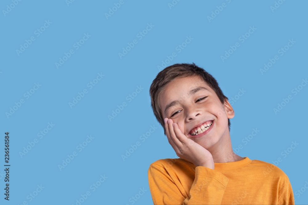 Close up emotional portrait of a boy wearing a yellow  on blue background in studio with copy space.He holds the cheek with his hand and laughs at something
