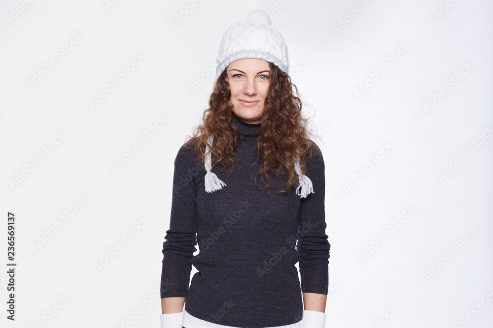 Portrait of attractive blue eyed young Caucasian woman with curly hair, wearing knitted cap with pompon, roll-neck grey sweater, looking confident at camera, being ready for cold times, posing indoors Portrait of attractive blue eyed young Caucasian woman with curly hair, wearing knitted cap with pompon, roll-neck grey sweater, looking confident at camera, being ready for cold times, posing indoors