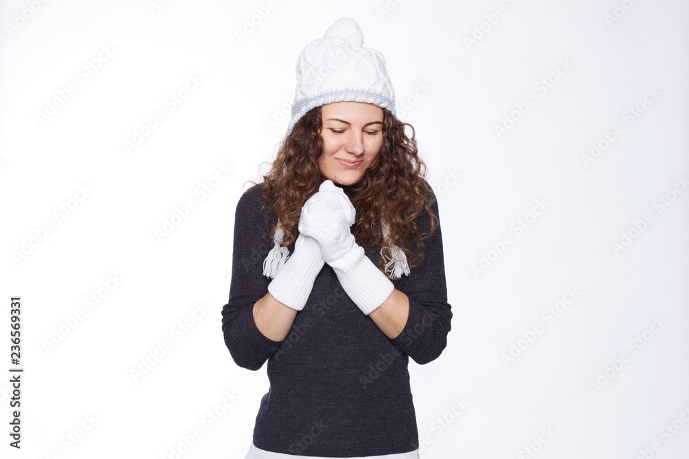 People, kindness and generosity concept. Warm dressed young comely woman in hat and mittens keeps hands on heart, expresses cordiality and friendliness, has charming smile, poses against studio wall. People, kindness and generosity concept. Warm dressed young comely woman in hat and mittens keeps hands on heart, expresses cordiality and friendliness, has charming smile, poses against studio wall.