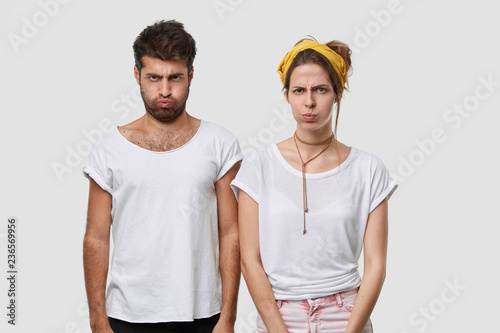 Gloomy sullen woman and man blow cheek, dont speak to each other after quarrel, have disagreement, insulted by bad words, stand shoulder to shoulder indoor, wear mockup white casual t shirt.