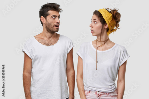 Suprised female and male discuss something unbelievable, look with widely opened eyes, have surprising facial expression, wear casual white t shirt, model indoor. People and reaction concept