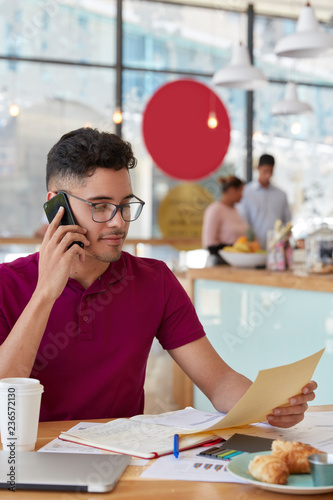 Intelligent creative young male designer has phone conversation, holds cellular, focused in paper document, compares figures, makes notes in diary, models against cafe interior. Vertical shot photo