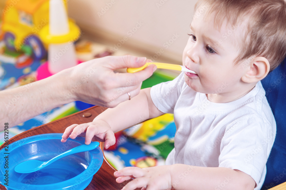 Little baby boy eating on a chair in the kitchen. Mom feeds holding in hand a spoon of porridge Little baby boy eating on a chair in the kitchen. Mom feeds holding in hand a spoon of porridge