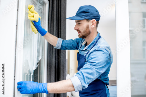 Man as a professional cleaner in blue uniform washing window with cotton wiper indoors
