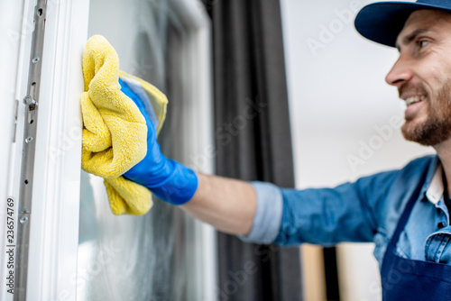 Man as a professional cleaner in blue uniform washing window with cotton wiper indoors