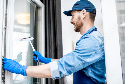 Man as a professional cleaner in blue uniform washing window with rubber brush indoors