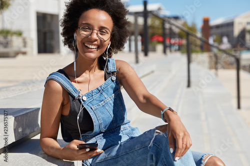 Glad black relaxed young woman listens favourite music or radio broadcasting, looks positively at camera, laughs happily, wears casual clothes, transparent glasses, models outdoor at street.