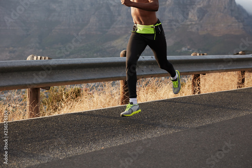 Unrecognizable dark skinned man fast runner sprints outside, runs in nature landscape background, leads healthy lifestyle, wears comfortable sportshoes. Sport exercise concept. Image with copy space photo