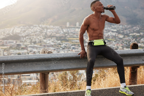 Shot of male runner drinks water after mountain running, feels thirsty, exercises outdoor early in morning, wears sportsclothes, rests at road banner, copy space on left. Sport, weariness, drinking photo