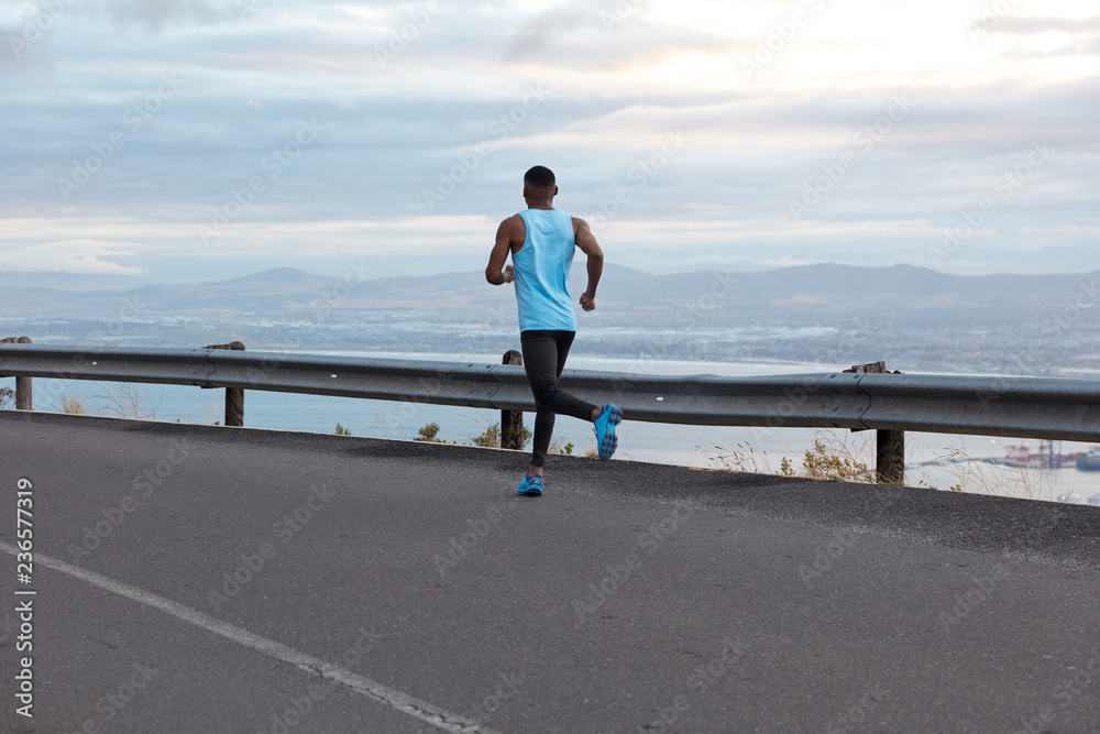 Back view of active young man with muscular body shape, dressed in sportswear, covers destination along road against blue sky in morning, enjoys fresh air, freedom and summer time. Sport concept.