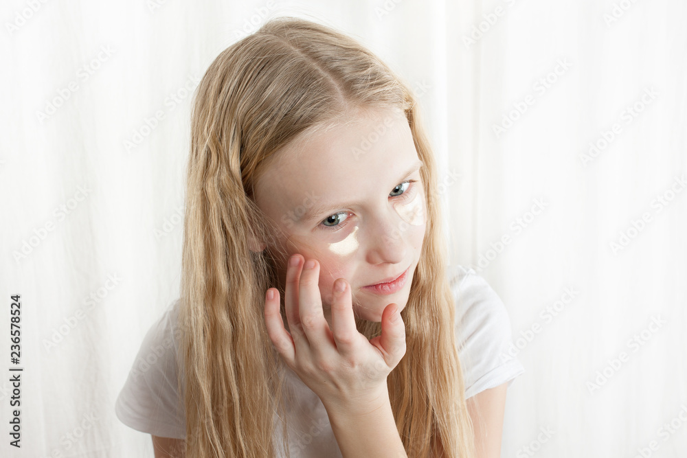 Young girl with curly and blonde hair trying on her mother's moisturizing patches from edema and tyred eyes. Isolated photo on white background. Young girl with curly and blonde hair trying on her mother's moisturizing patches from edema and tyred eyes. Isolated photo on white background.