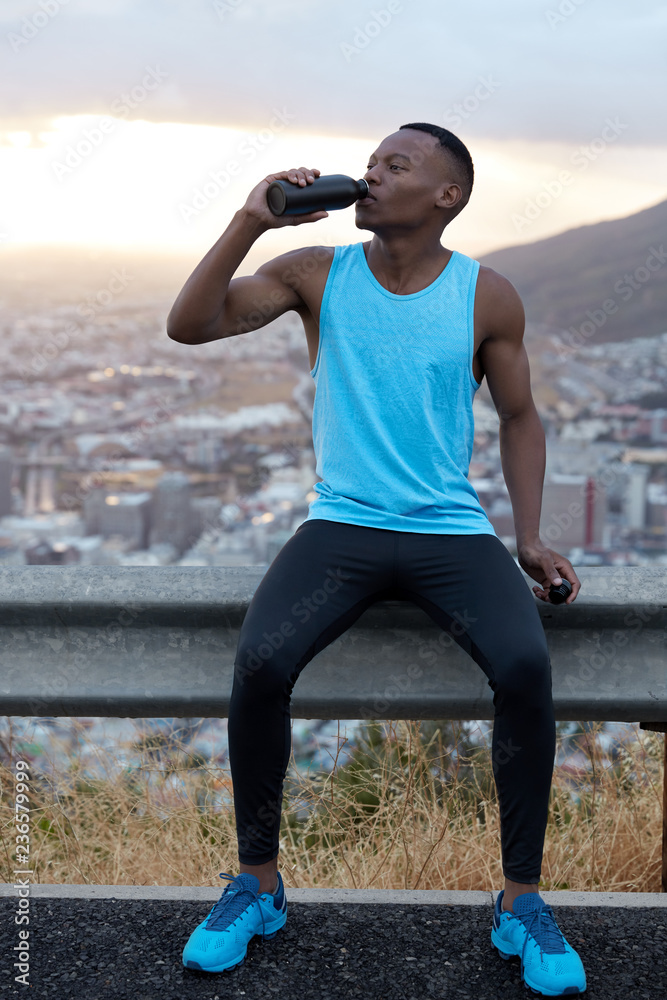 Vertical shot of handsome African American man in sportswear drinks water  after fitness training, refreshes with beverage, poses over mountain hill,  feels fatigue. Sport and rejuvenation concept Stock Photo | Adobe Stock