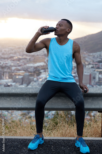 Fototapeta Naklejka Na Ścianę i Meble -  Vertical shot of handsome African American man in sportswear drinks water after fitness training, refreshes with beverage, poses over mountain hill, feels fatigue. Sport and rejuvenation concept