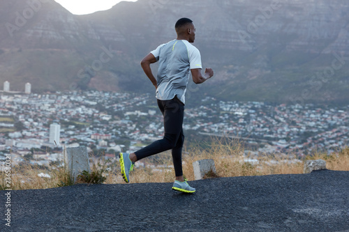 Photo of sportsman in casual t shirt, black leggings and trainers, runs quickly along mountain road, being fast jogger, poses against nice landscape in countryside, being self determined and strong photo