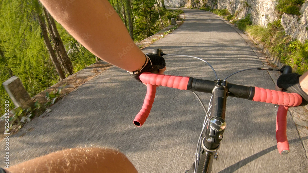 Naklejka premium POV: Pedalling a road bicycle up a steep asphalt road in the sunny mountains.