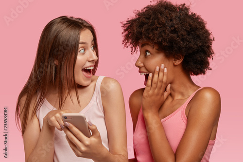 Surprised joyful interracial friends look happily at screen of cell phone, read something unbelievable, pose together against pink background, make shopping online. Ethnicity and technology.