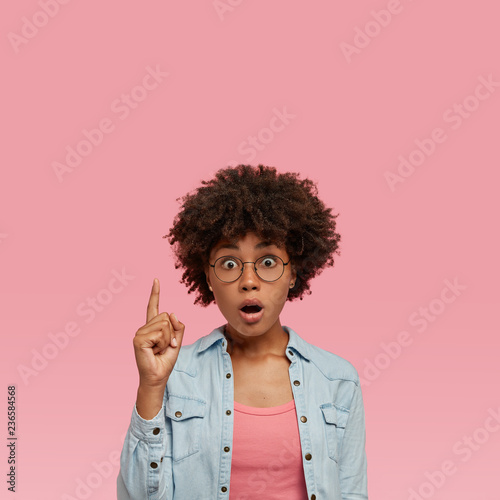 Shocked beautiful hipster teenager with dark skin, opened mouth, dressed in fashionable denim shirt, points with index finger upwards, being impressed to notice hole in ceiling, models indoor