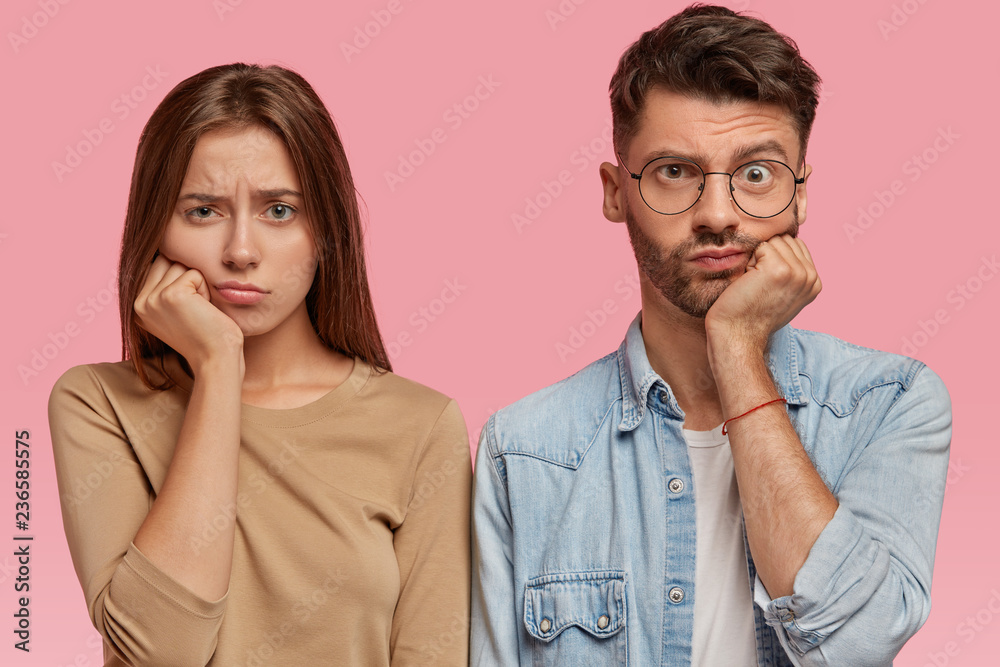 Bored girl and boy keep hand under chin, look with sadness directly at camera, feel dissatisfied, wait for something for long time, stand against pink background. People and facial expressions concept Bored girl and boy keep hand under chin, look with sadness directly at camera, feel dissatisfied, wait for something for long time, stand against pink background. People and facial expressions concept