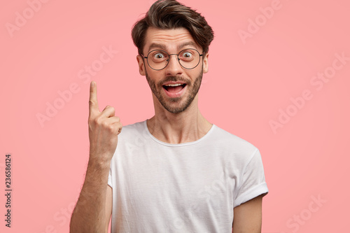 Surprised hipster with trendy haircut, has intriguing shocked gaze, points with index finger upwards, wears casual white t shirt and spectacles, isolated over pink background. Advertisement.