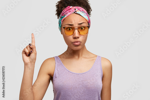 Studio shot of indignant puzzled Afro American lady looks with worried expression at camera  points with index finger upwards  dressed in casual outfit  trendy sunglasses  isolated over white wall