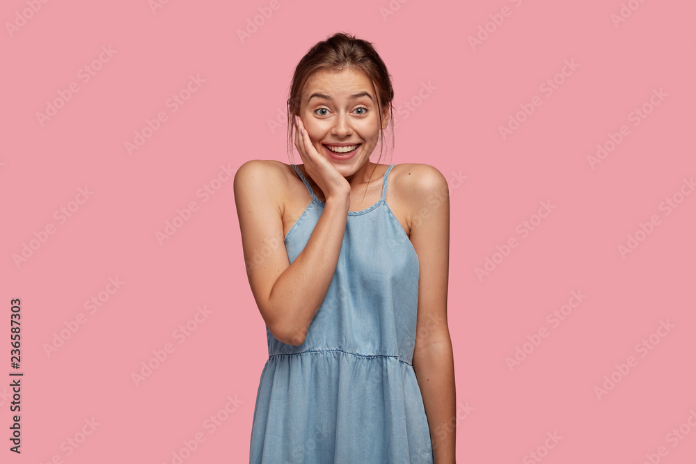 Joyful pretty dark haired woman has toothy smile, keeps hand on cheek, dressed in denim dress, models against pink background, happy to recieve compliment from husband, expresses positiveness Joyful pretty dark haired woman has toothy smile, keeps hand on cheek, dressed in denim dress, models against pink background, happy to recieve compliment from husband, expresses positiveness