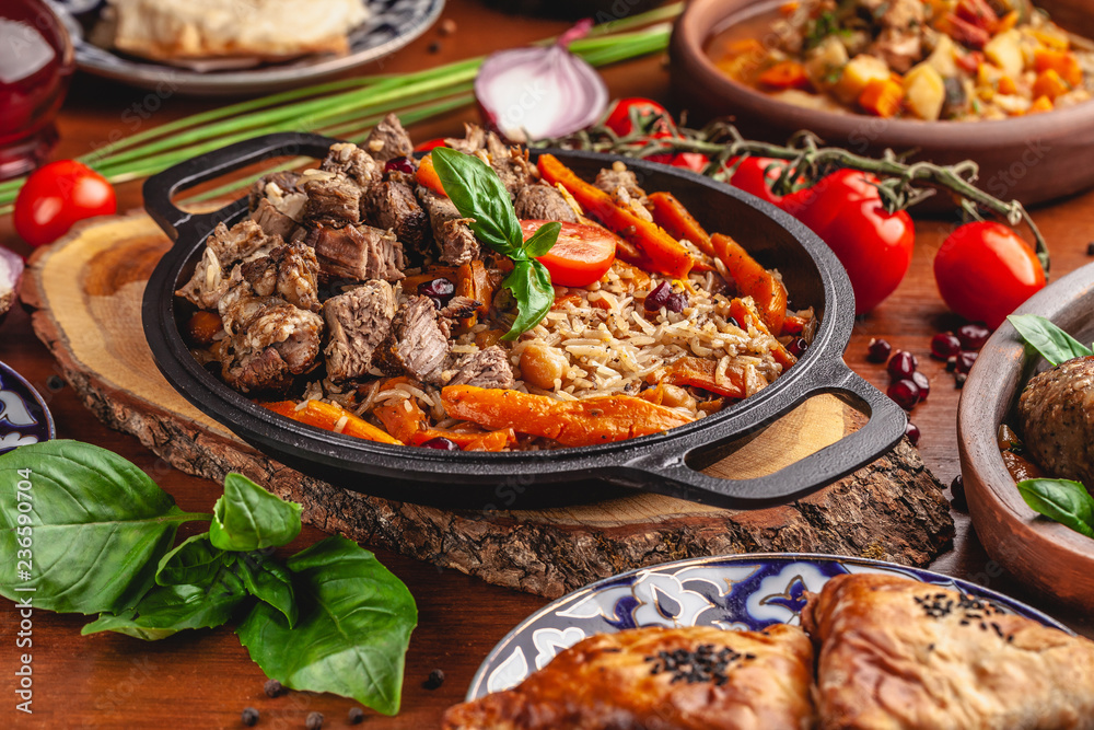 The concept of Uzbek, oriental cuisine. Pilaf in a cast-iron pan, with pieces of lamb lies on a wooden table. Festive table set.