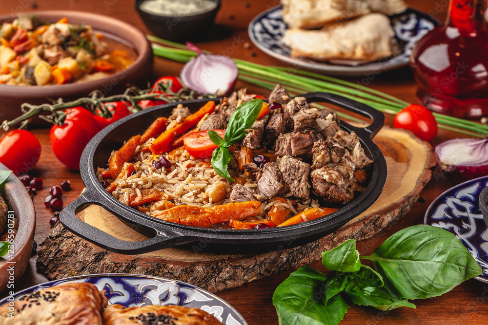 The concept of Uzbek, oriental cuisine. Pilaf in a cast-iron pan, with pieces of lamb lies on a wooden table. Festive table set.
