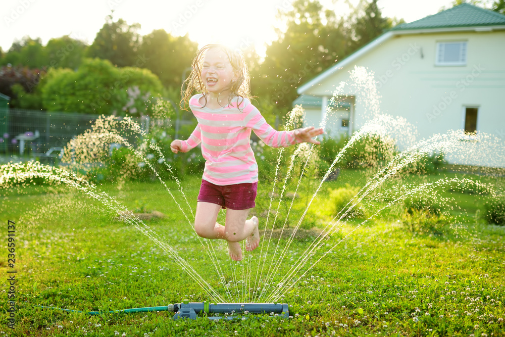 Adorable little girl playing with a sprinkler in a backyard on sunny summer day. Cute child having fun with water outdoors. - obrazy, fototapety, plakaty 