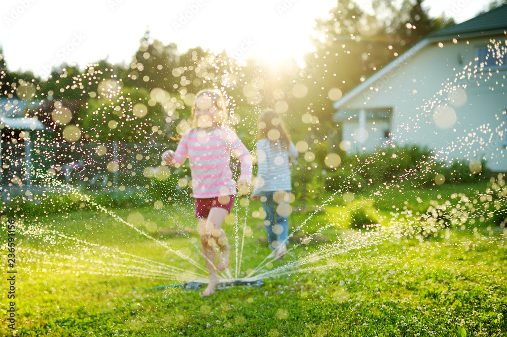 Adorable little girls playing with a sprinkler in a backyard on sunny summer day. Cute children having fun with water outdoors. - obrazy, fototapety, plakaty 