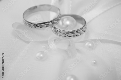 Two gold wedding engagement rings on a white pad with a bow and beads, close-up. Black and white tone