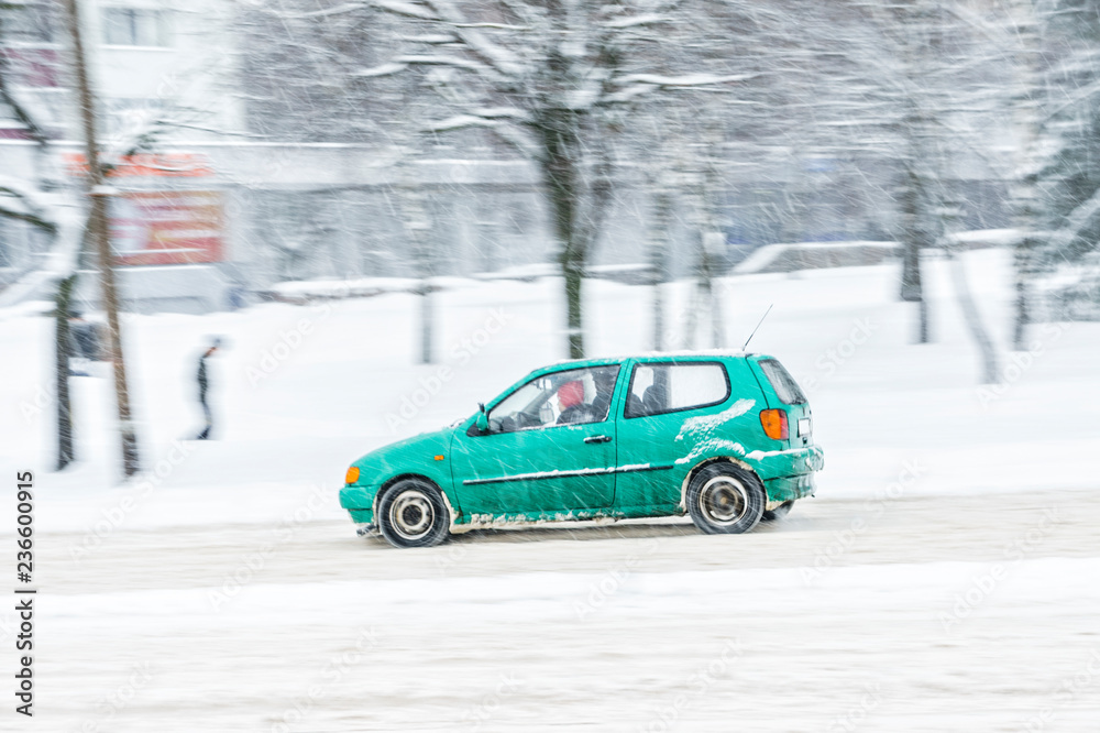 Driving in snow. Motion in blur small green car in heavy snowfall in city road. Abstract blur winter weather background